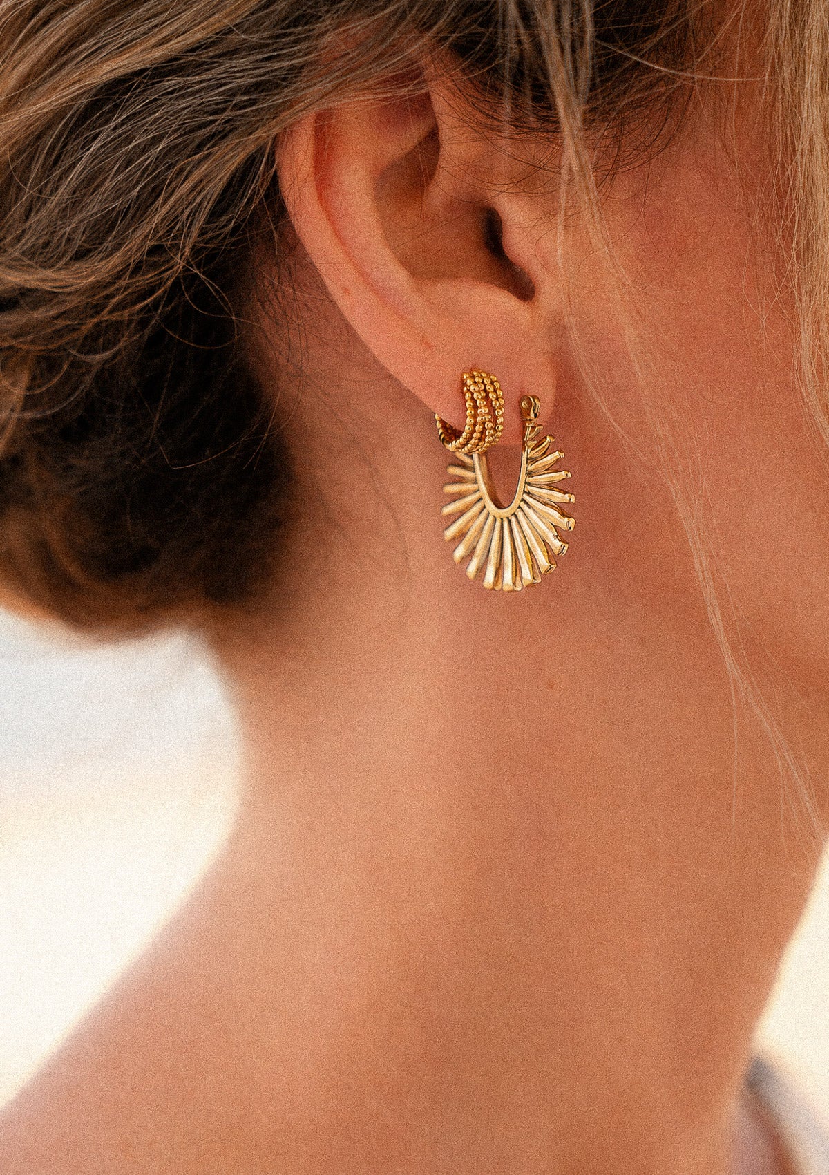 Boucles d'oreilles Soleil Rayonnant portées à l'oreille, demi-cercle doré en éventail à rayons de soleil, en accumulation avec créole perlée dorée cheveux blonds au vent – Namaka collection Solaire