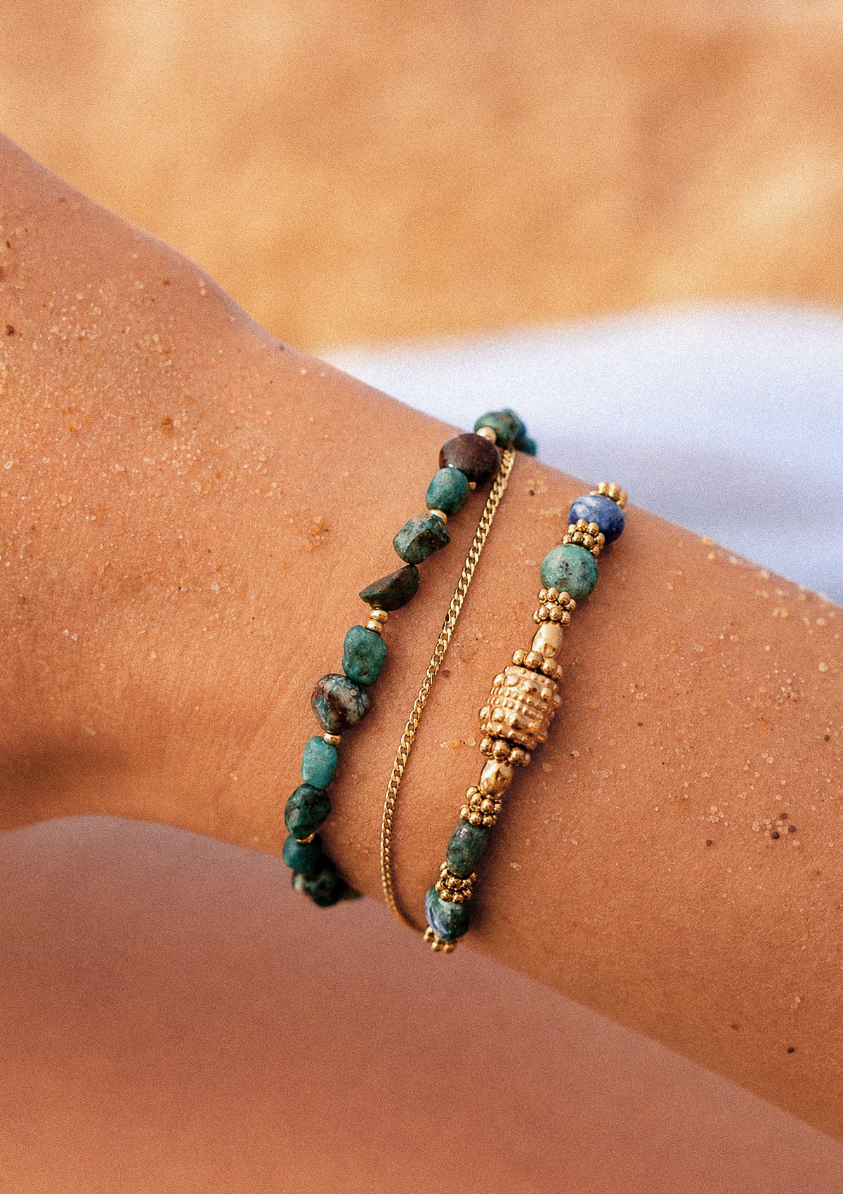 Bracelet Azurite Ligne d'horizon porté en accumulation avec chaîne fine dorée et bracelet perles dorées, poignet ensablé sur plage au soleil couchant – Namaka collection Solaire