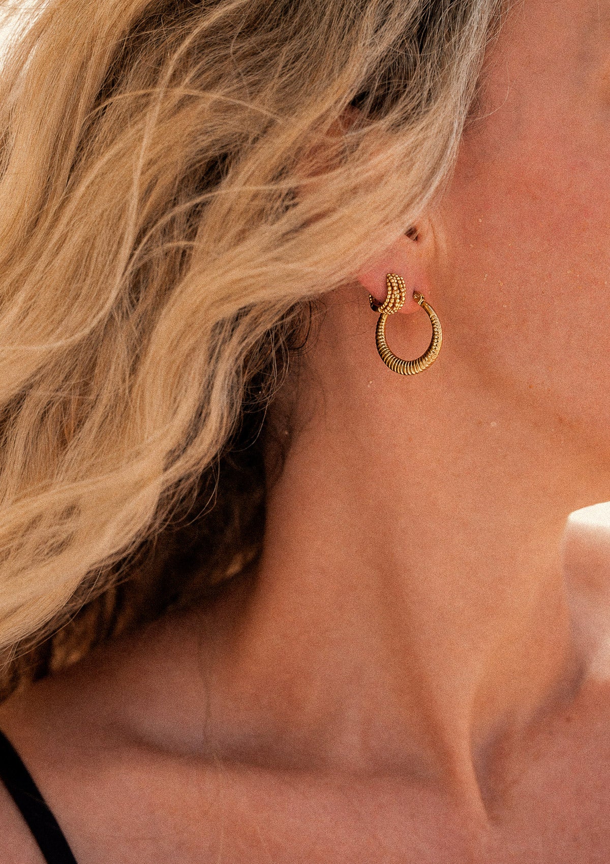 Créoles Gravées Rayons Bruts, anneau doré à stries en rayons de soleil, portées à l'oreille en accumulation avec créole perlée dorée cheveux blonds au vent – Namaka collection Solaire