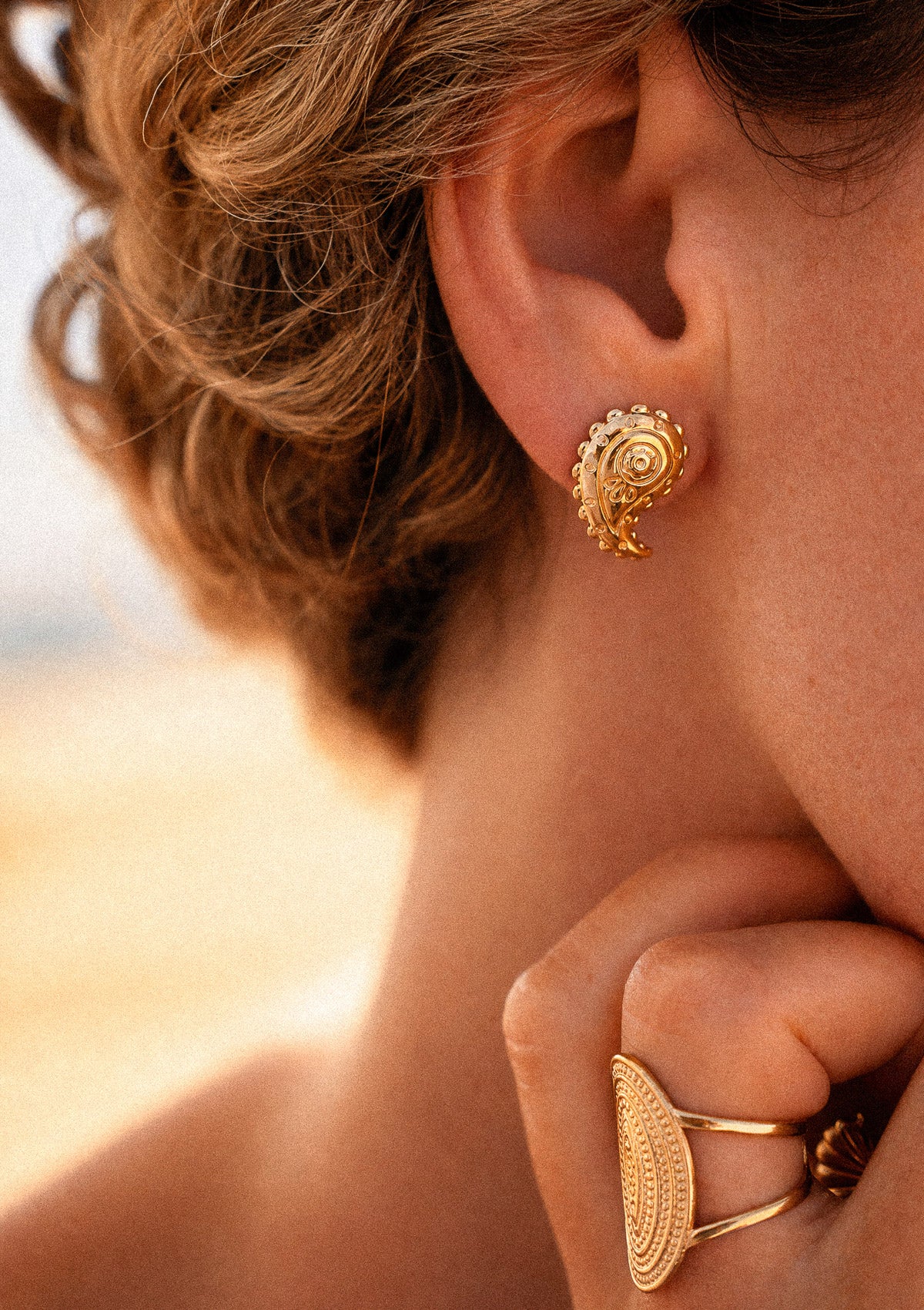 Boucles d'oreilles Trésor Nomade portées à l'oreille, motif ethnique paisley doré finement gravé, en accumulation avec bague ovale dorée en lumière solaire – Namaka collection Solaire
