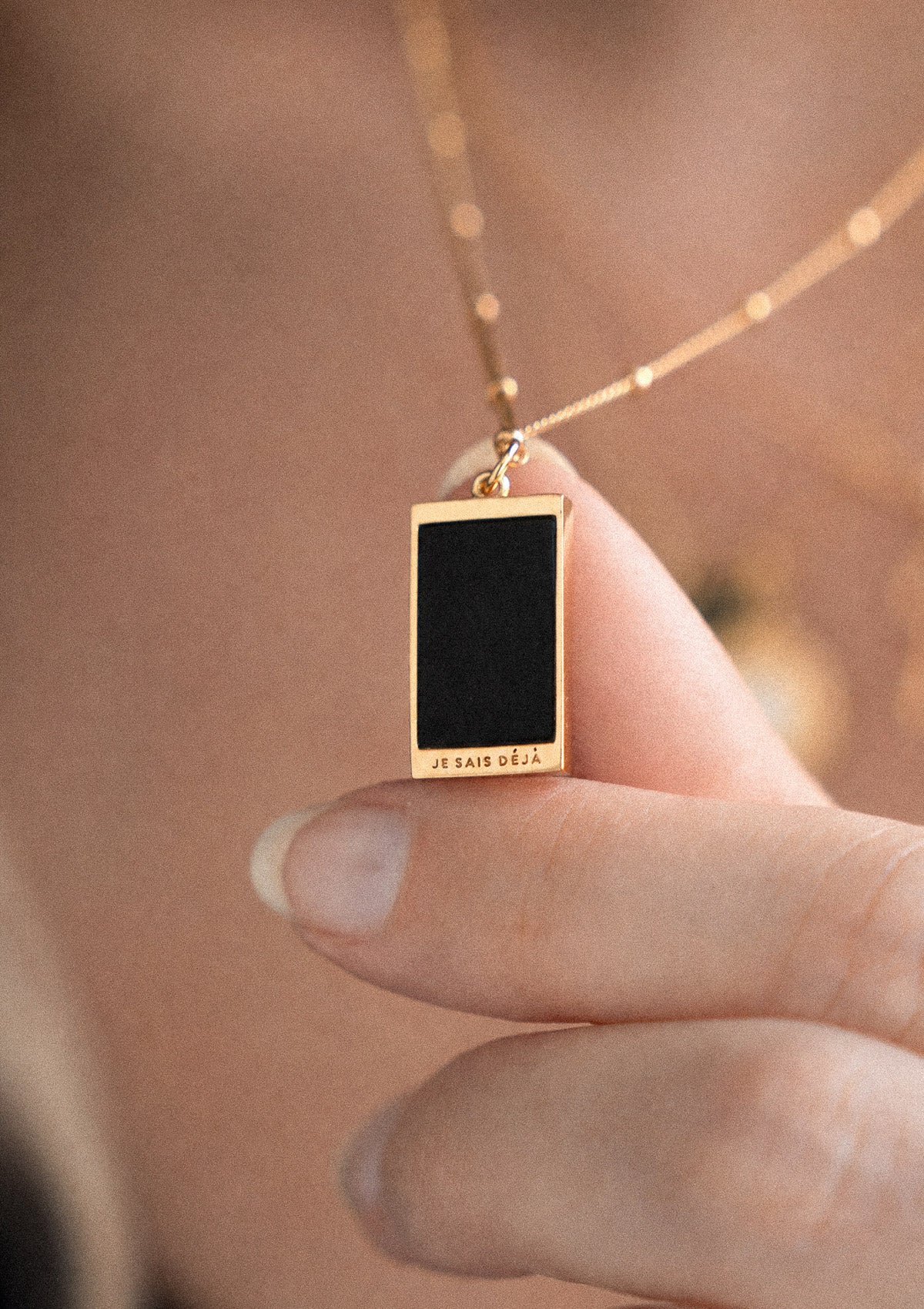 Revers pendentif tarot or 18 carats plaque Onyx Noir mat gravure JE SAIS DÉJÀ tenu entre pouce et index, chaîne à billes dorée
