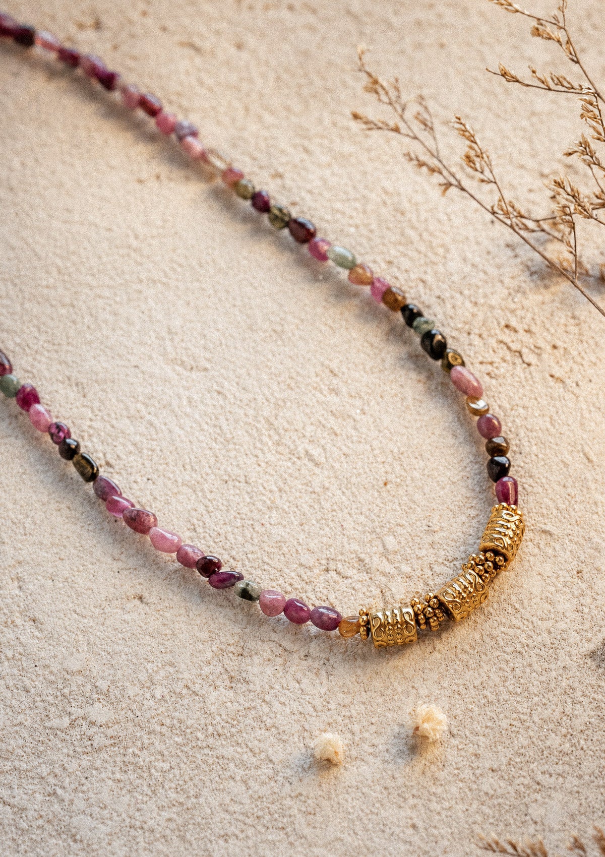 Collier Harmonie Vivante posé sur sable en bord de plage, ras-de-cou Tourmaline multicolore et détails perlés dorés en lumière chaude – Namaka collection Solaire, assemblé en France