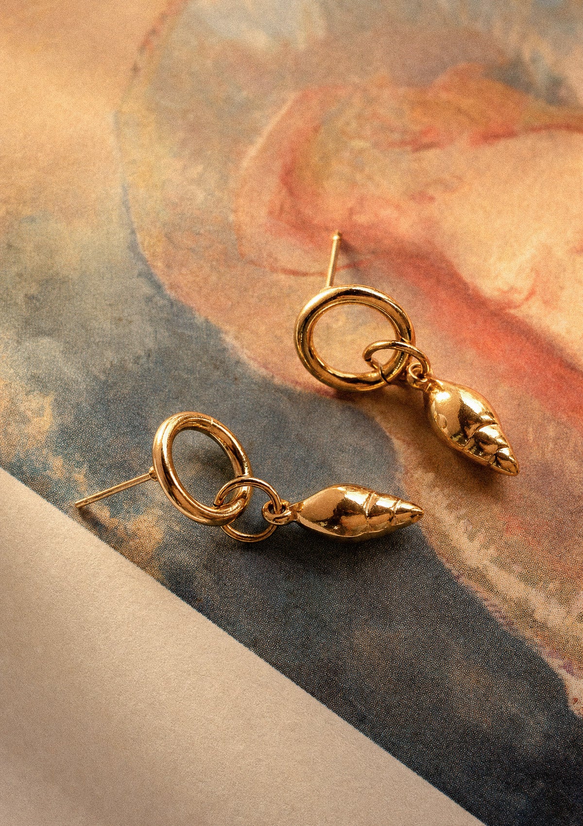 Boucles d'oreilles coquillage Sel d'or acier inoxydable doré, présentées sur fond aquarelle rose et bleu – bijoux d'été Namaka collection Solaire, assemblées en France