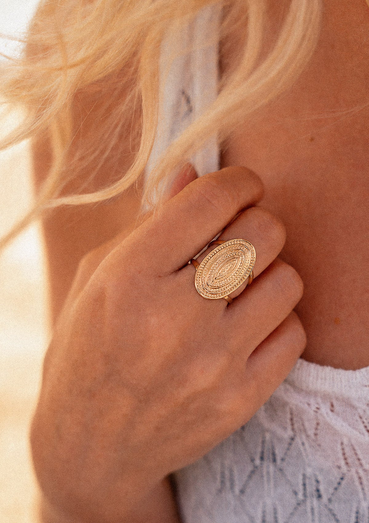 Bague Ovale Astre Intérieur portée à la main, motif ovale doré aux ellipses perlées concentriques en gros plan lumière dorée de plage – Namaka collection Solaire