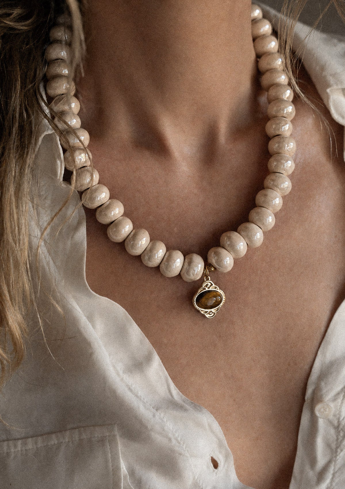 Collier ras-de-cou grosses perles céramique crème et pendentif Œil de Tigre porté sur chemise blanche, pierre de courage et de confiance