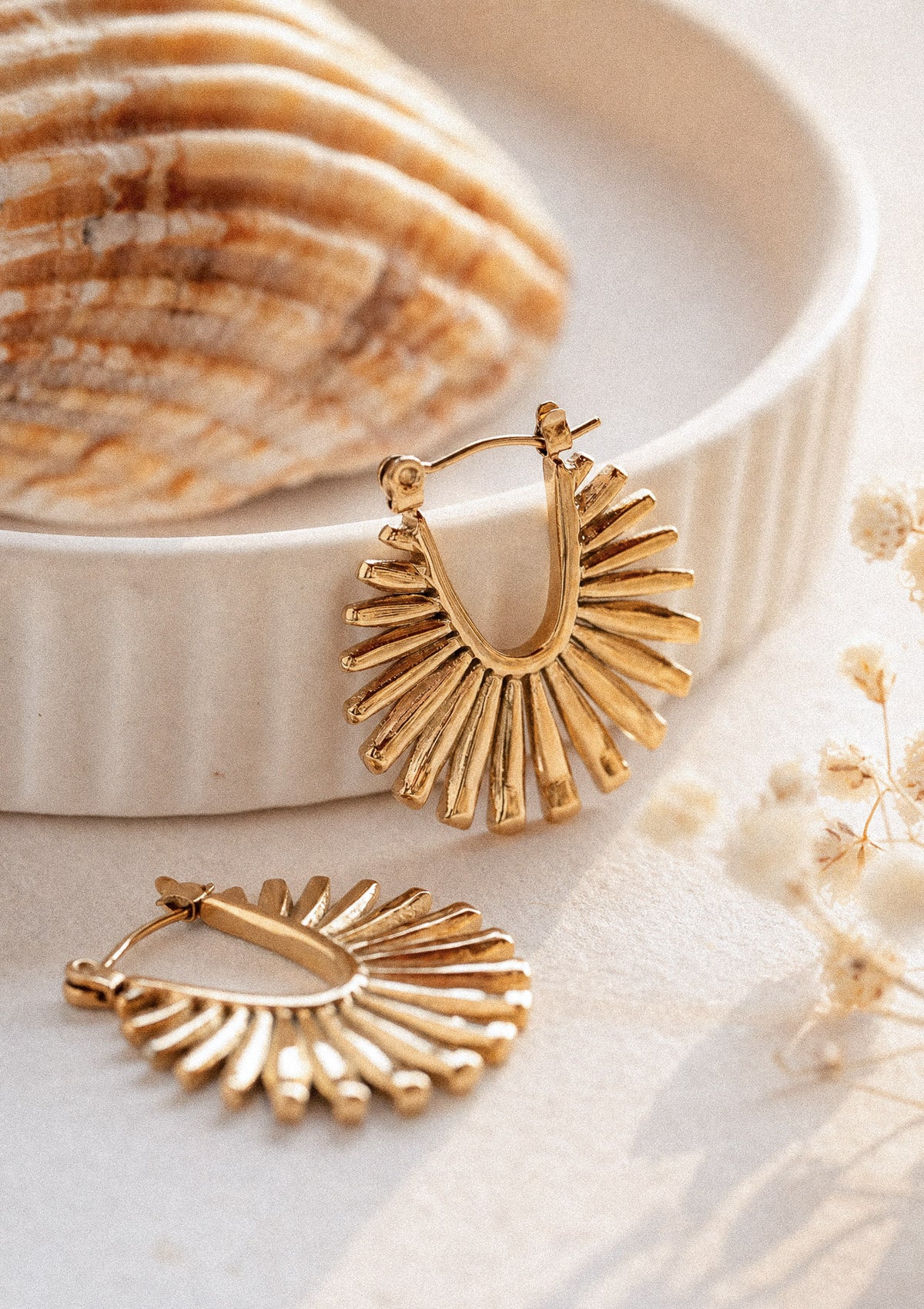 Boucles d'oreilles Soleil Rayonnant packshot, deux créoles en éventail doré massif rayonnant, dans une coupelle blanche près d'un coquillage – acier inoxydable doré Namaka collection Solaire