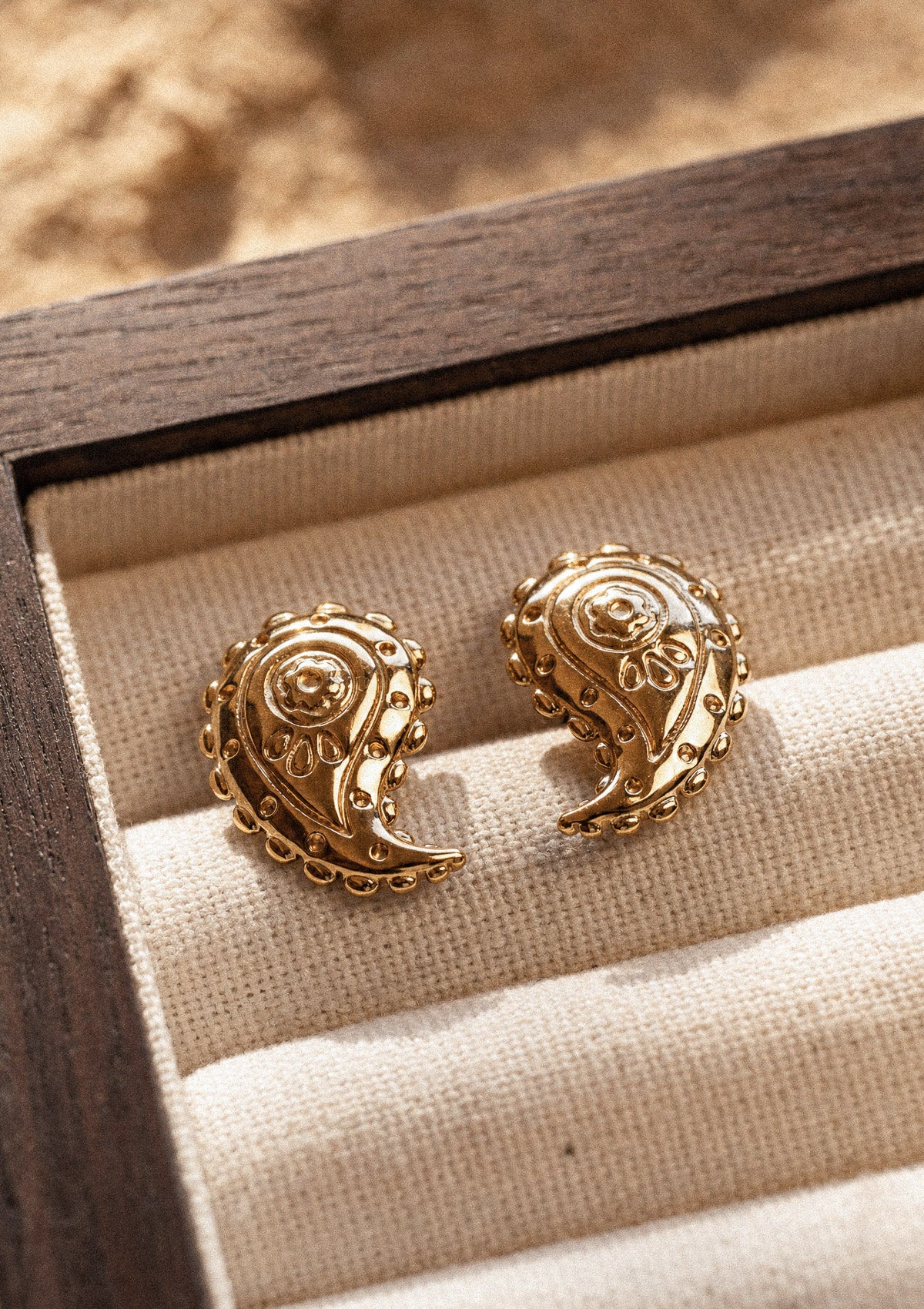 Puces d'oreilles Trésor Nomade au motif paisley cachemire doré ciselé, dans une boîte à bijoux en bois sur sable – acier inoxydable doré Namaka collection Solaire