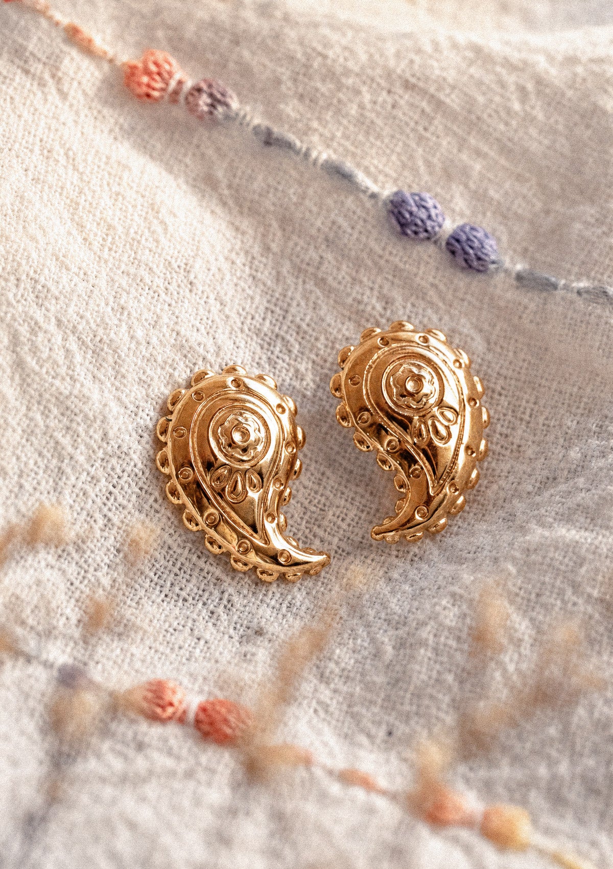 Puces d'oreilles paisley dorées Trésor Nomade, motif cachemire ethnique ciselé, posées sur tissu tissé écru à pompons brodés – acier inoxydable doré Namaka collection Solaire