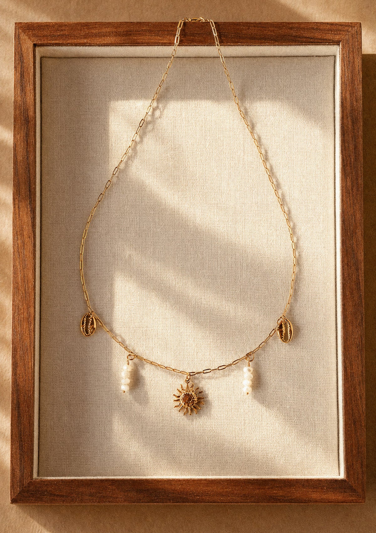 Collier Rivage d'Été packshot dans cadre bois, chaîne dorée avec pendentif soleil rayonnant, coquillages cauri dorés et perles de Nacre – Namaka collection Solaire, assemblé en France