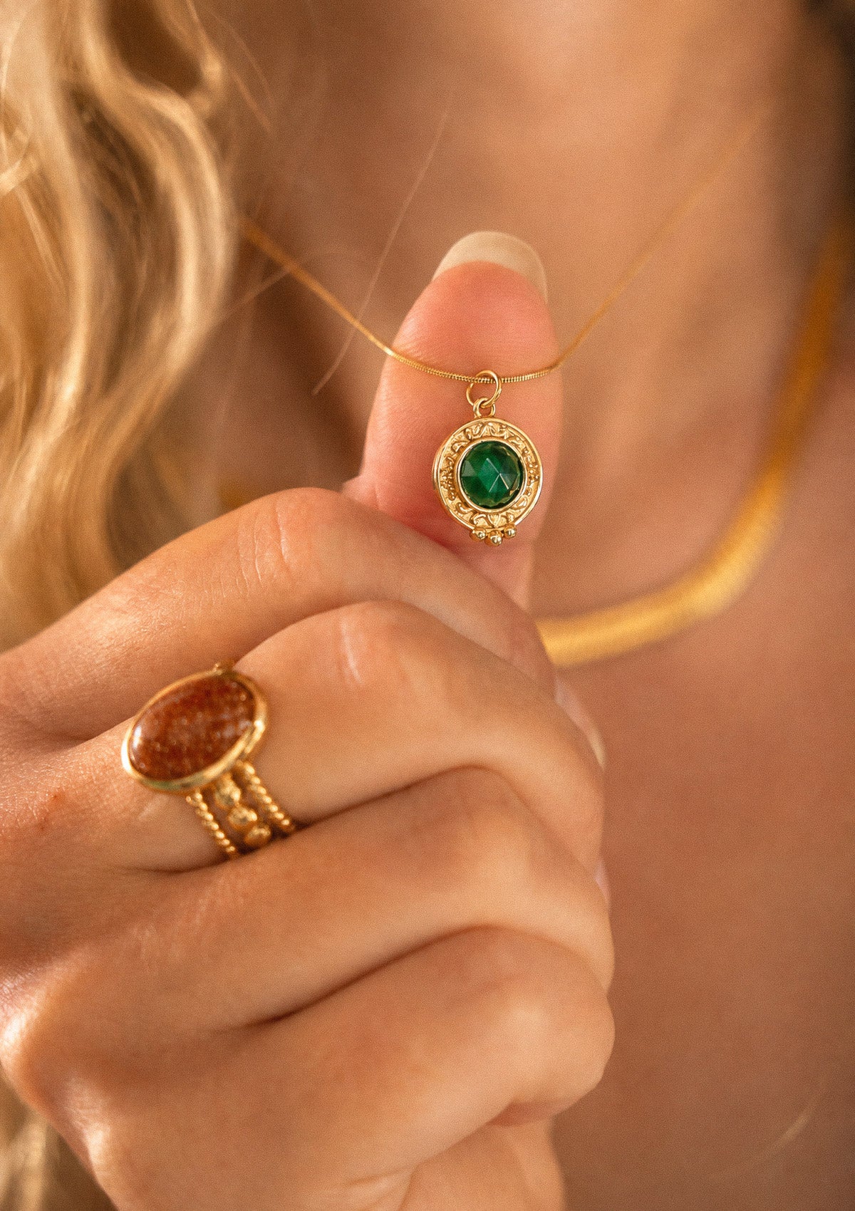 Pendentif Malachite tenu en main avec la bague Pierre de Sable au doigt, chaînes dorées en arrière-plan signé Namaka