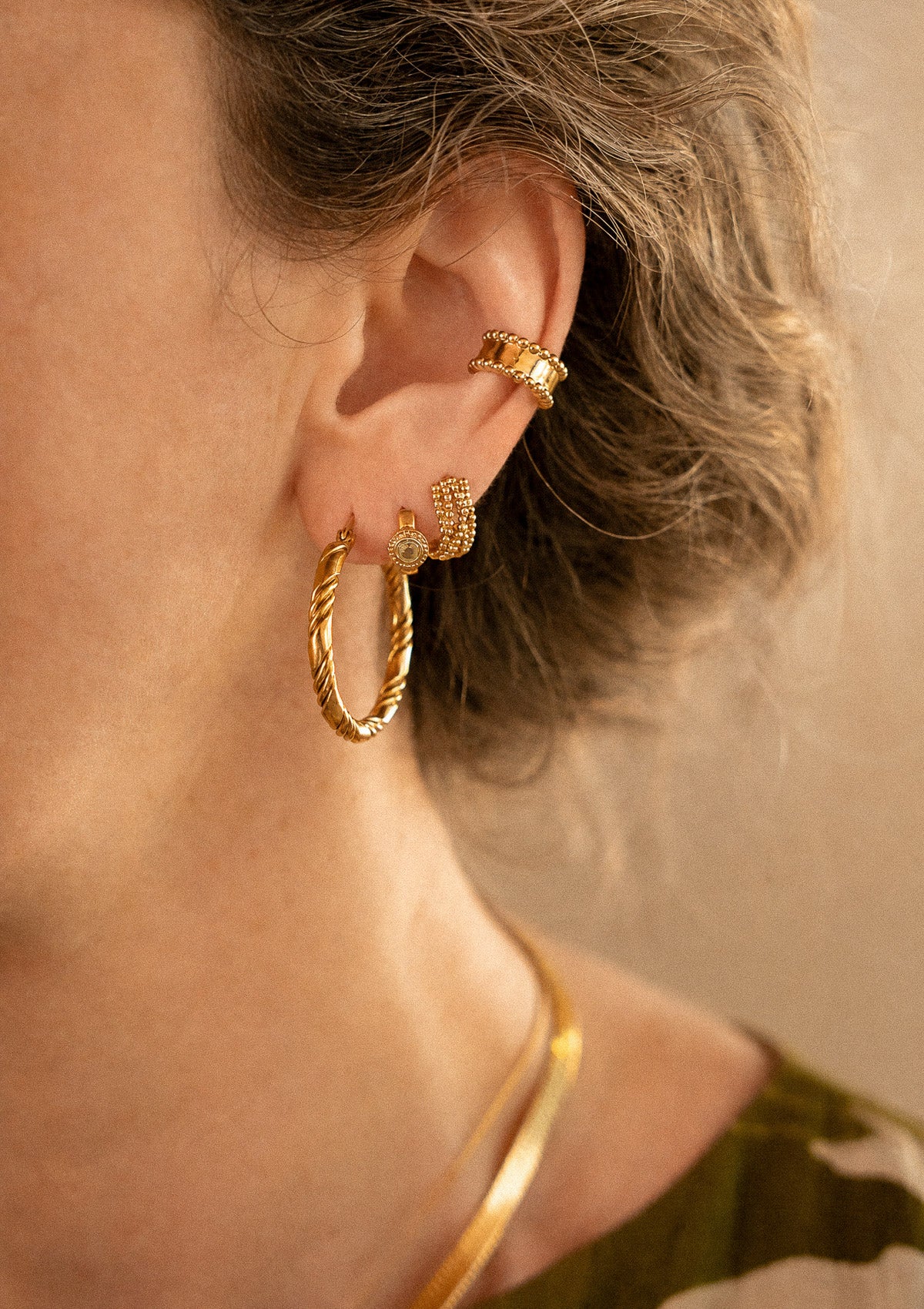 Détail des grandes créoles torsadées dorées portées de profil avec mini créole Péridot et ear cuff perlé Namaka